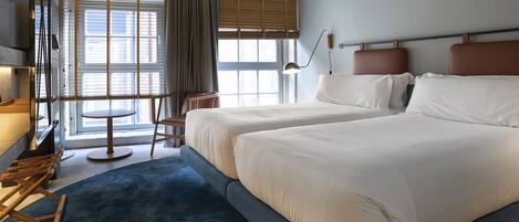Habitación de hotel con dos camas, un escritorio, una silla, una lámpara y vistas al exterior a través de amplias ventanas.