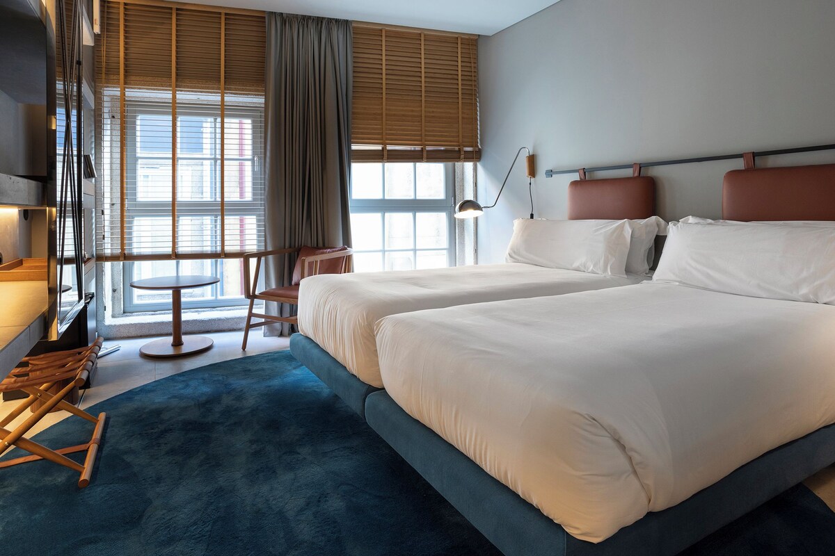 A hotel room with two beds, a desk, a chair, a lamp, and a view of the city through large windows.