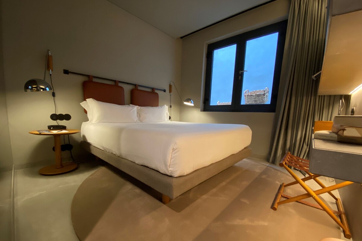 A hotel room with a bed, bedside table, lamp, and a view of the city through a window.