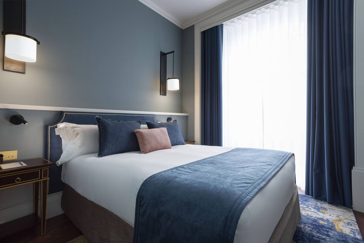 A neatly made bed with a blue blanket and pillows, a bedside table with a lamp, and a window with curtains.