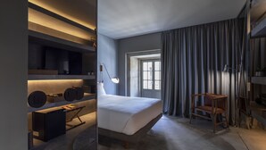 A modern bedroom with a large bed, bedside tables, a desk, and a chair. There is a window with curtains and a view of the outside. - ICON Duplo Ribeira (Porto)
