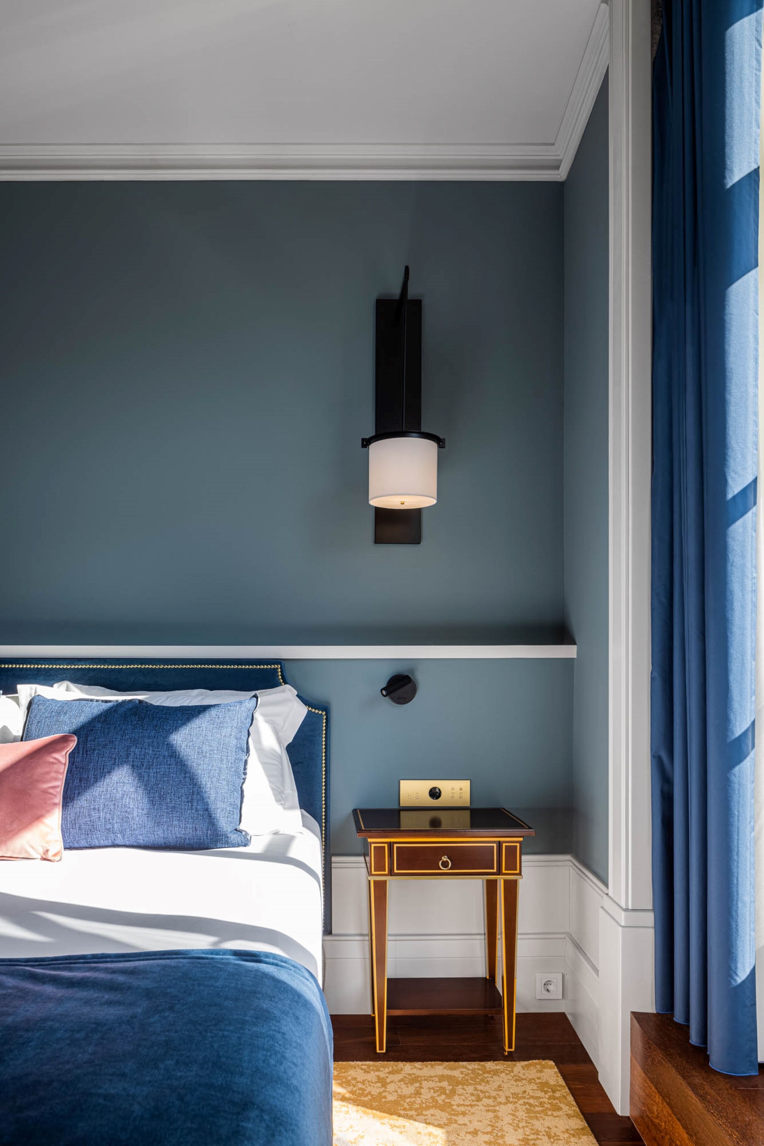A bedroom with a bed, a nightstand, a wall-mounted lamp, and a window with curtains.