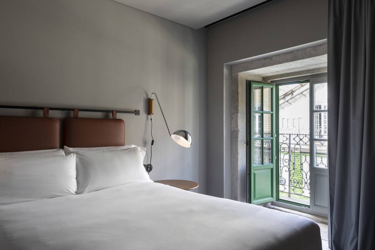 A modern hotel room with a large bed, a bedside table, a lamp, and a window with a view of a balcony.