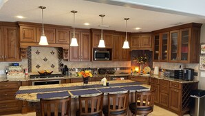 A kitchen with wooden cabinets, a central island with bar stools, pendant lighting, and a microwave. - Eastpoint on the  Bay - private Beach, pool, sunsets (Eastpoint)