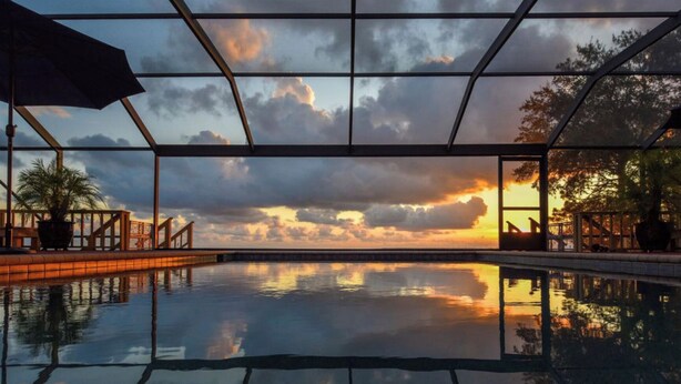 A swimming pool with a sunset view through a glass enclosure, surrounded by a wooden deck and potted plants. - Eastpoint on the  Bay - private Beach, pool, sunsets (Eastpoint)