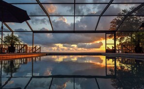 A swimming pool with a sunset view through a glass enclosure, surrounded by a wooden deck and potted plants. - Eastpoint on the  Bay - private Beach, pool, sunsets (Eastpoint)