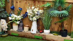 A wooden fence with various potted plants and flowers, including white and blue flowers, and a welcome sign. - Greenham Rise (Stoke-sub-Hamdon)