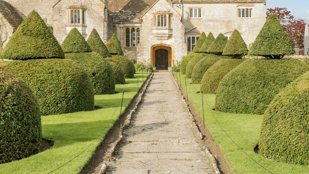 A stone building with a central entrance, surrounded by neatly trimmed hedges and a stone pathway leading to the entrance. - Greenham Rise (Stoke-sub-Hamdon)