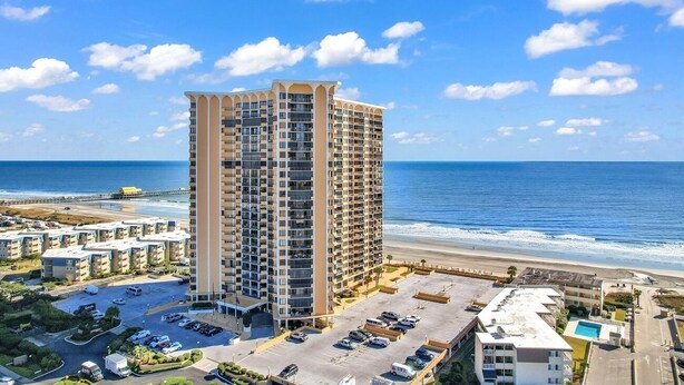 A high-rise hotel with multiple balconies, a parking lot, and a view of the beach and ocean.