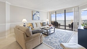 A spacious living room with a beige sofa, a coffee table, and a patterned rug. There is a balcony with outdoor furniture and a view of the city skyline.