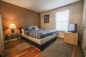 A bedroom with a bed, a nightstand, a lamp, a TV on a stand, and a window with blinds.