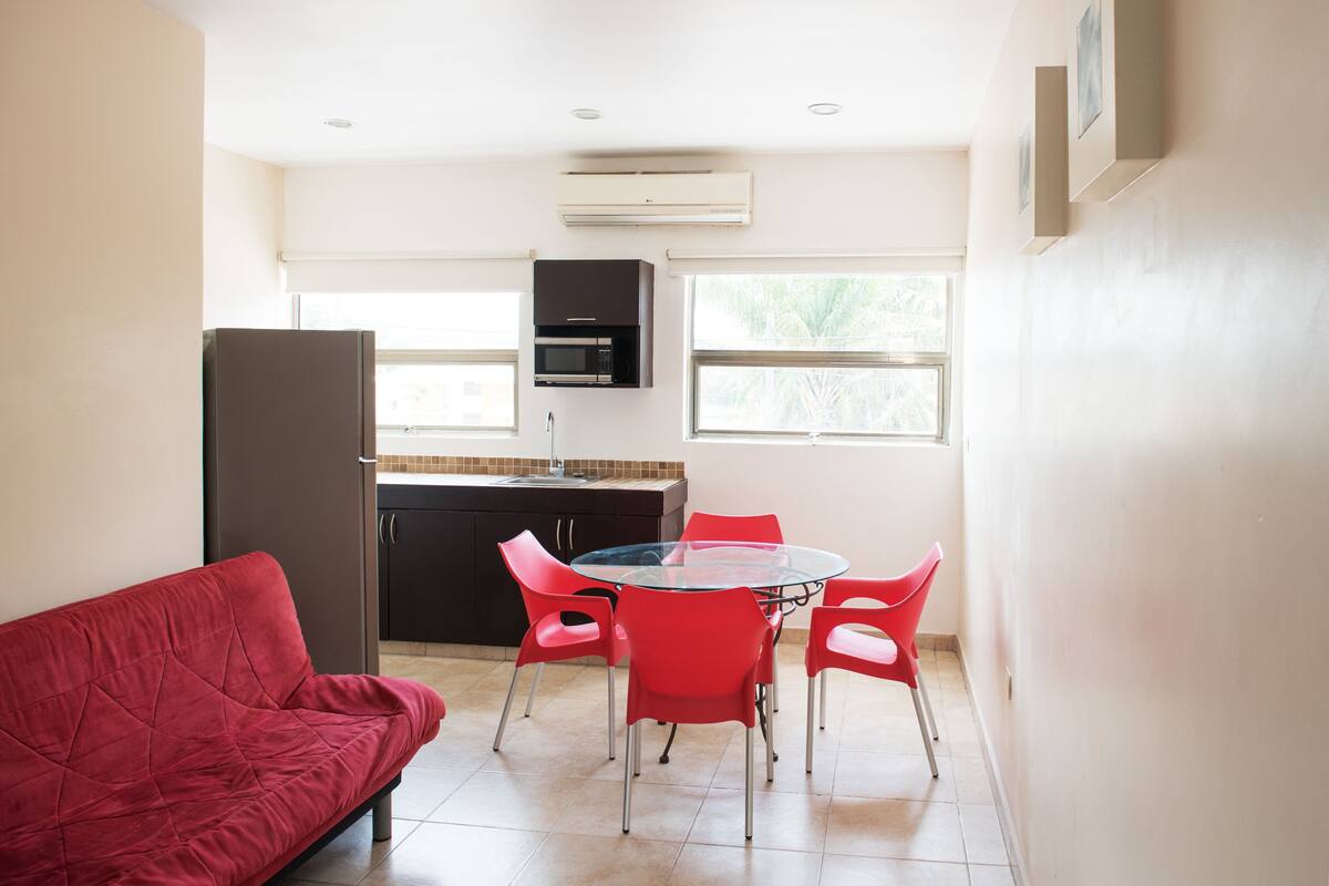 A compact kitchen with a dining area, including a red couch and red chairs around a glass table.