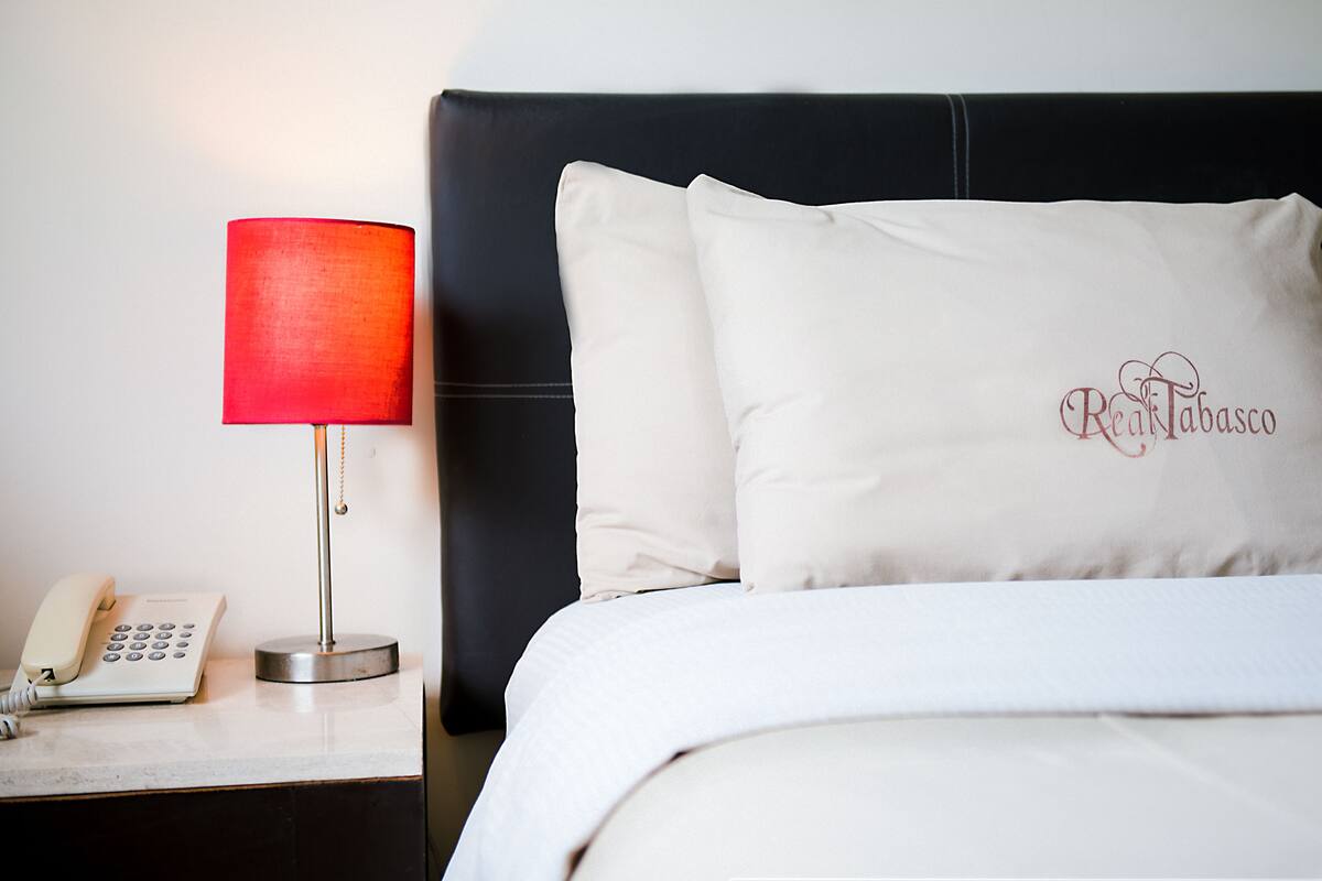 A hotel room with a bed, a nightstand, a red lamp, and a white pillow with "Real Tabasco" embroidered on it.