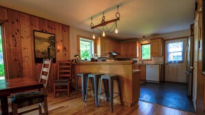A kitchen with wooden cabinets, a central island with stools, a hanging light fixture, and a window providing natural light. - BASE CAMP LA CONCEPTION (La Conception)