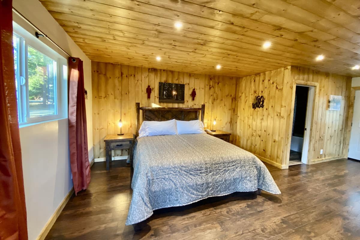 A wooden-paneled bedroom with a bed, bedside tables, and a door leading to another room.