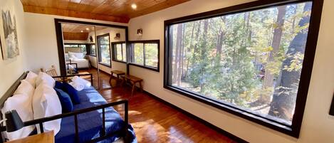 Una habitación con cama, suelo de madera, ventanas grandes y vistas a un bosque.