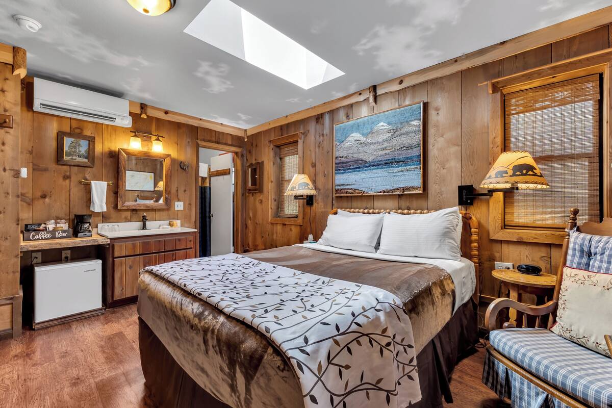 A cozy bedroom with a large bed, a wooden headboard, a small desk with a coffee station, a nightstand, and a window with a view of mountains.