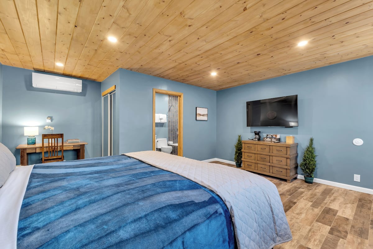 A bedroom with a bed, wooden ceiling, blue walls, a TV, a dresser, and a desk with a chair.