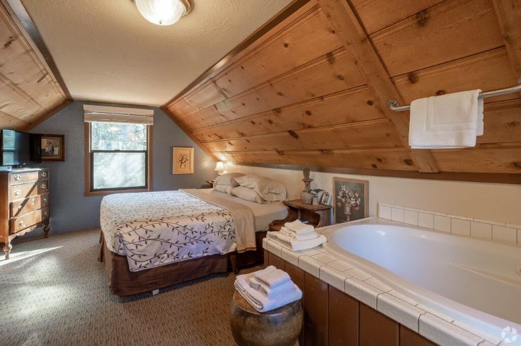A bedroom with a large bed, a wooden ceiling, a window, a dresser, and a bathtub.