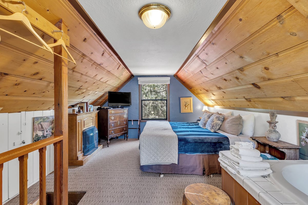 A cozy attic bedroom with a bed, a dresser, a vanity, and a window with a view of trees.