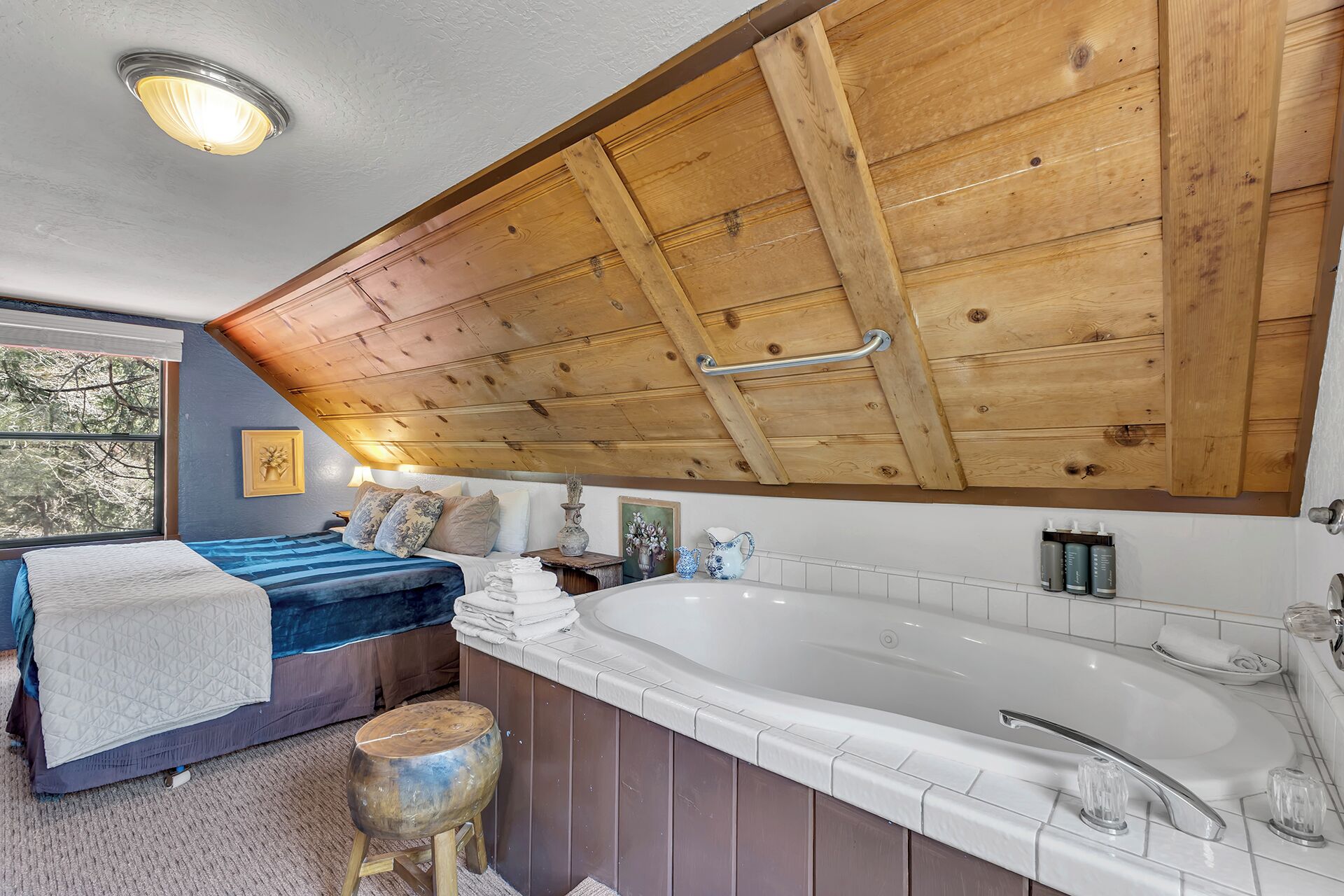 A bedroom with a large wooden ceiling, a bed with blue bedding, a wooden stool, a bathtub, and a window with a view of trees.
