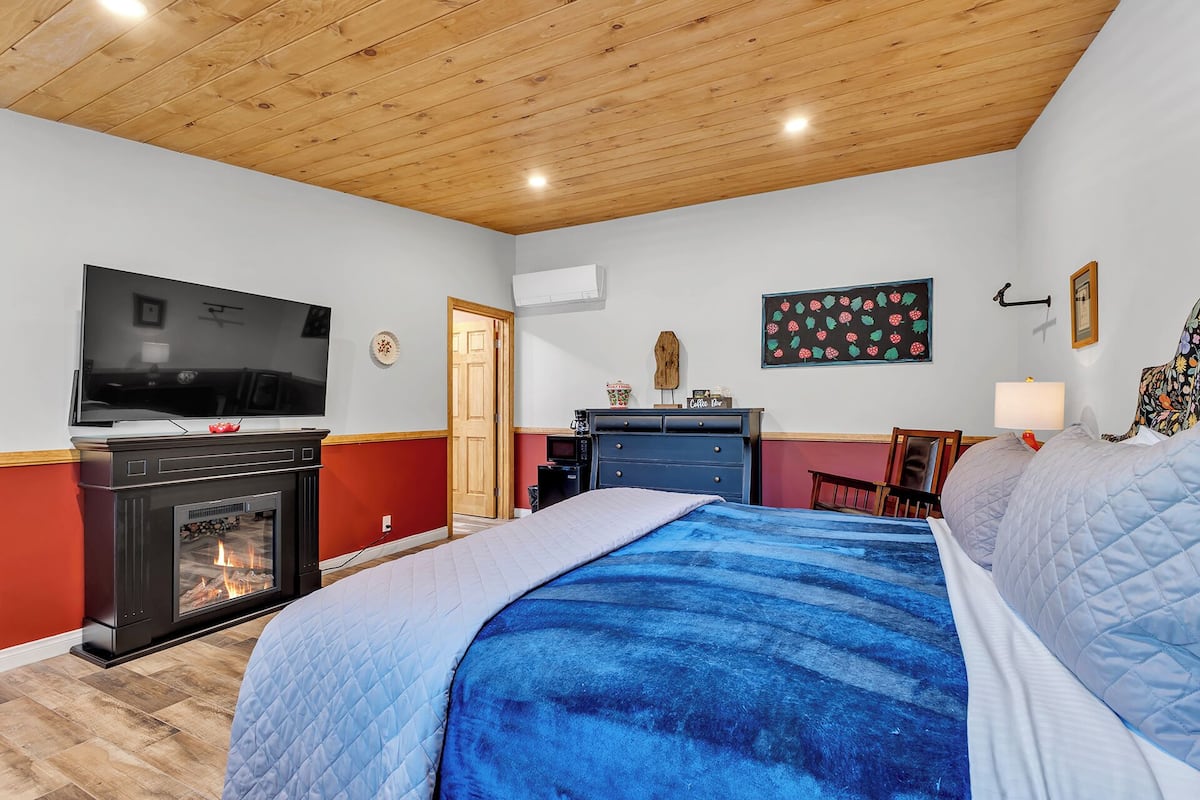 A bedroom with a bed, a television, a fireplace, a desk, and a wall-mounted lamp.