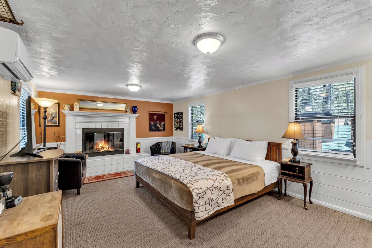 A bedroom with a fireplace, a bed with a patterned bedspread, a nightstand with a lamp, and a desk with a computer.