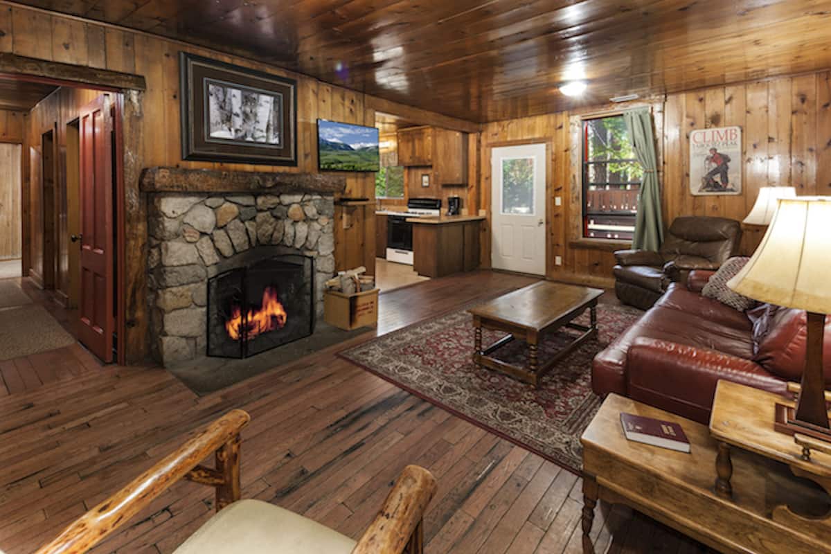 A cozy cabin interior with a stone fireplace, wooden floors, a leather sofa, and a dining area.
