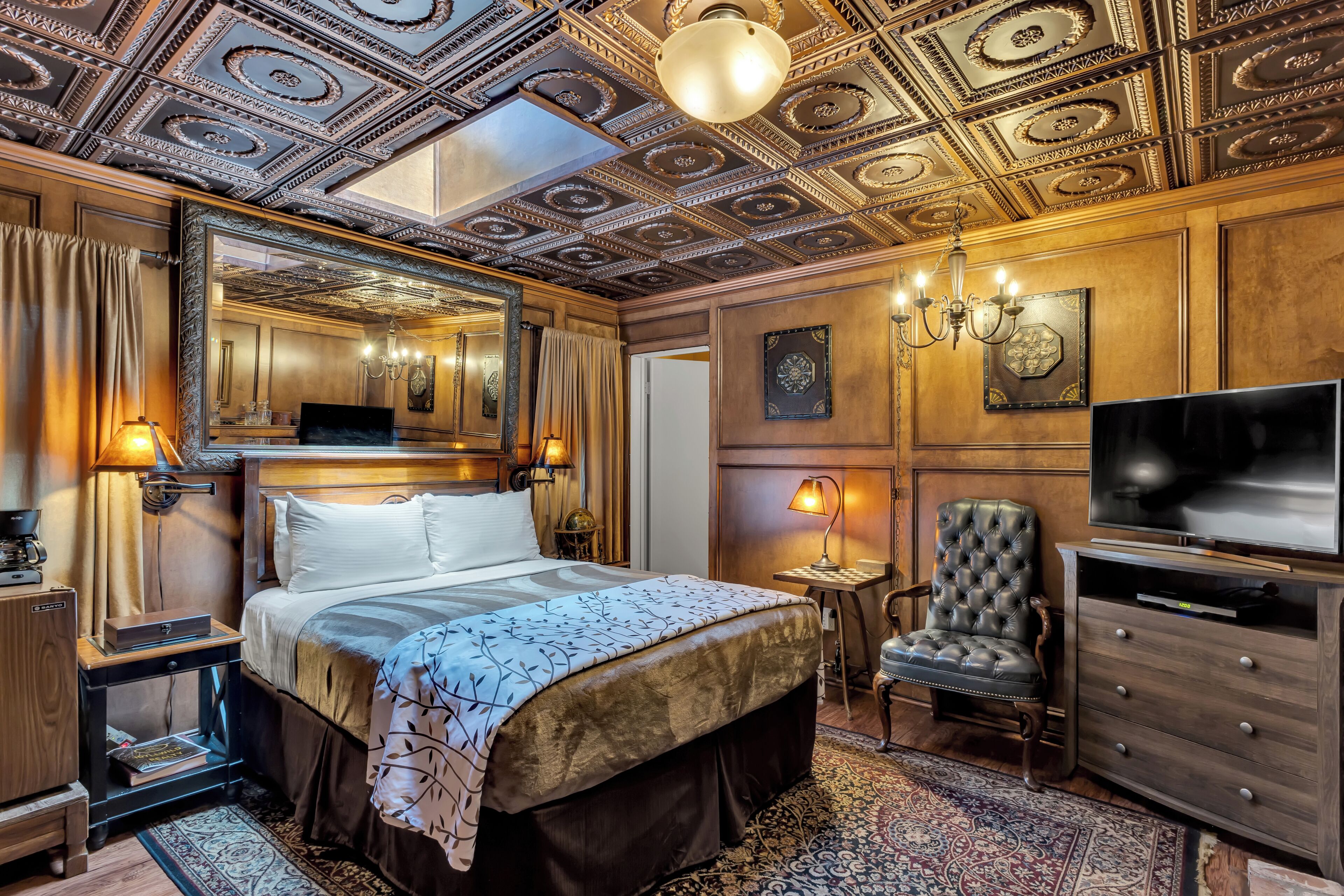 A luxurious bedroom with a large bed, a television, a leather armchair, and ornate ceiling details.