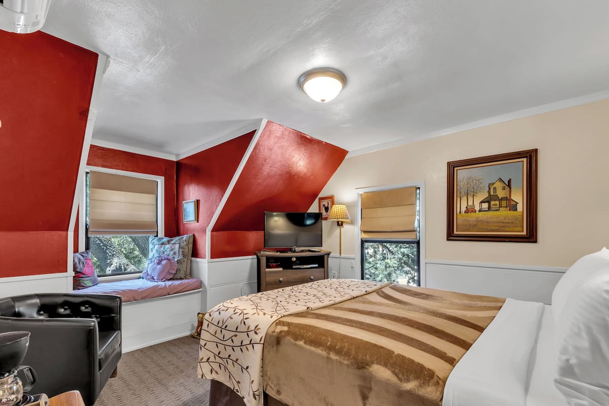 A bedroom with a sloped ceiling, a bed, a TV, a desk, and a window with a view of trees.