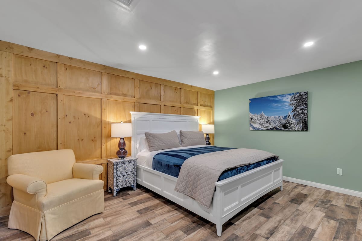 A bedroom with a wooden paneled wall, a bed with a white headboard, a beige armchair, and a nightstand with a lamp.