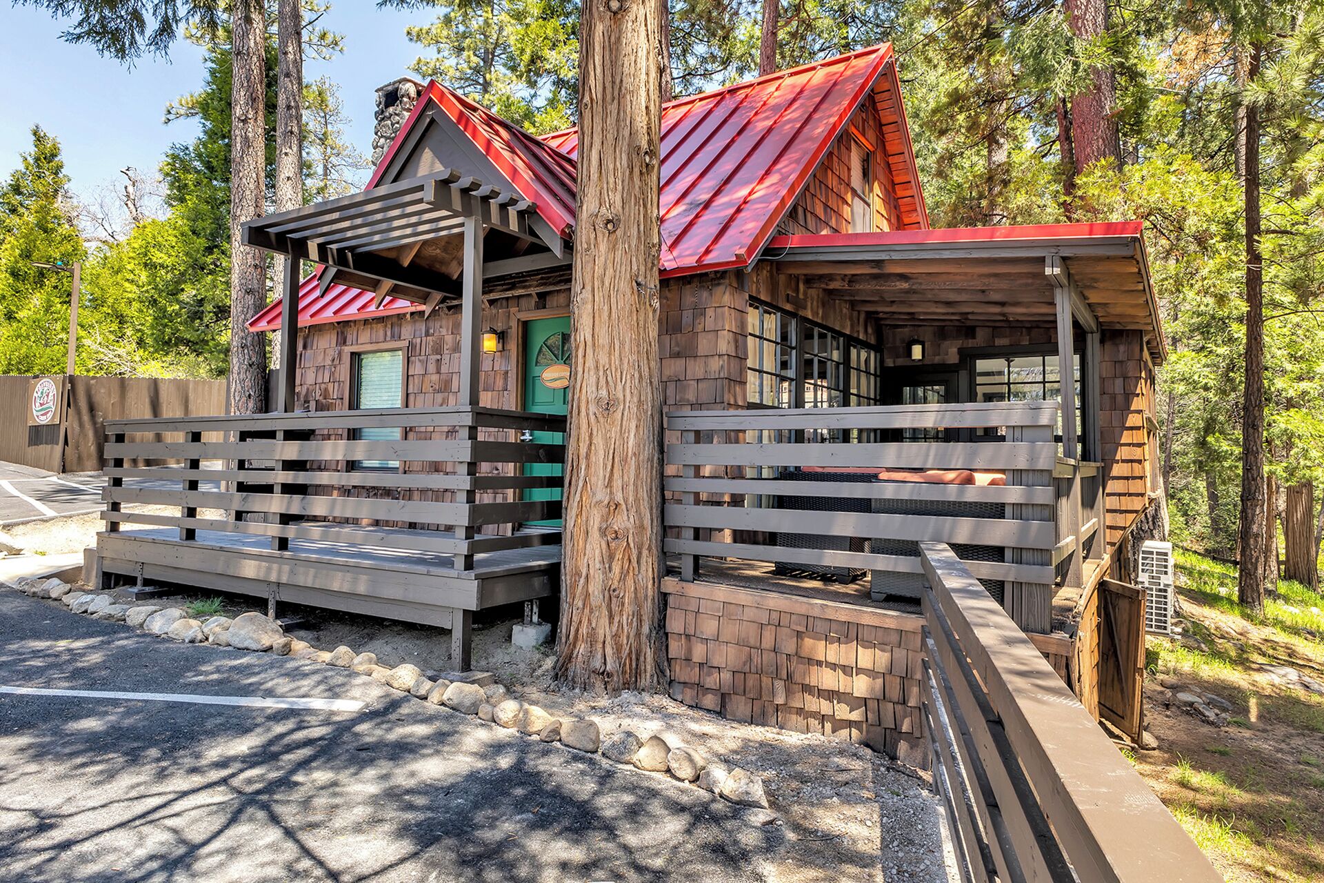 A rustic cabin with a wooden deck and a red roof, surrounded by trees.