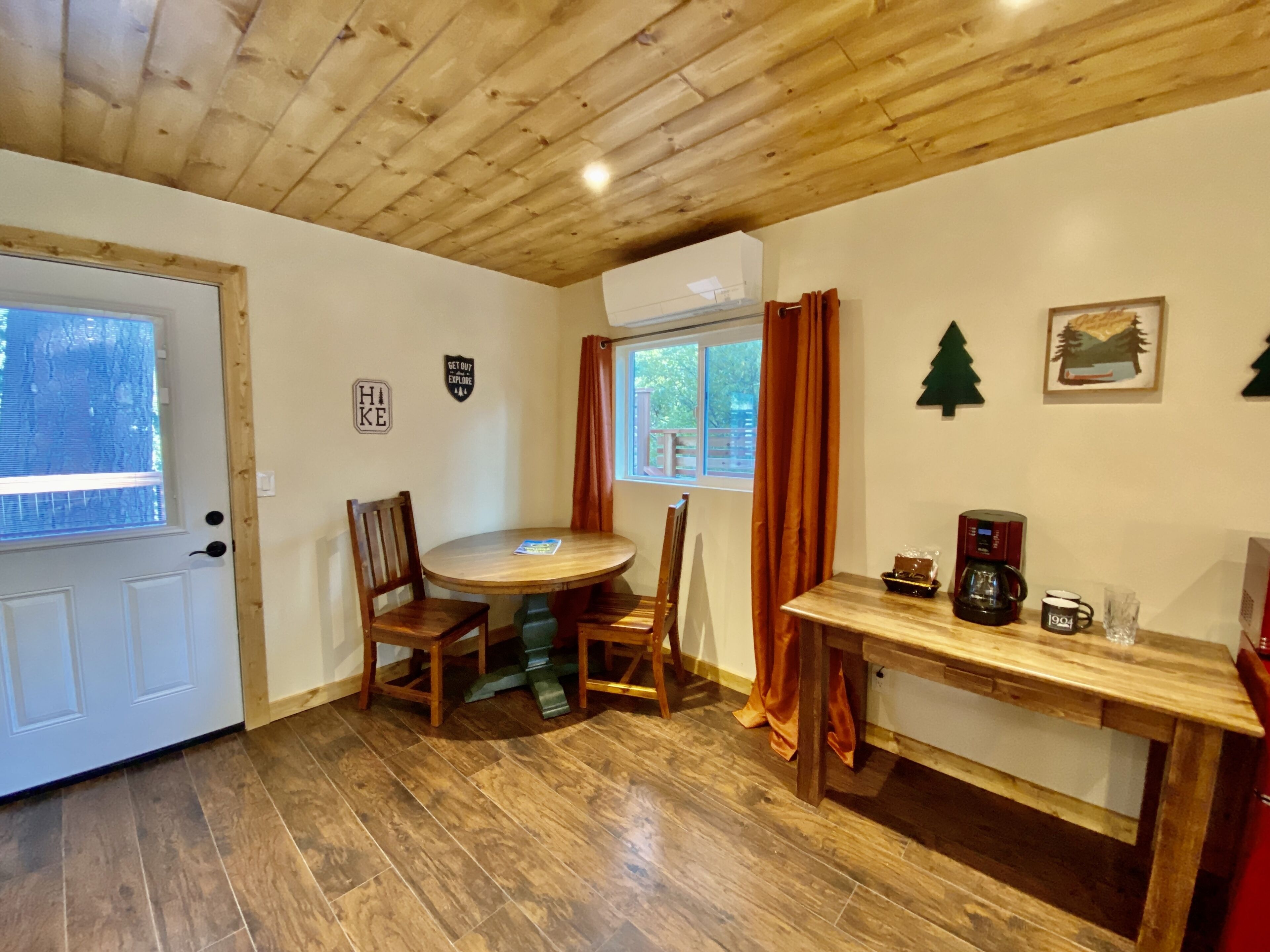 Una habitación con paneles de madera, una mesa de comedor, sillas, una cafetera y una ventana con cortinas.