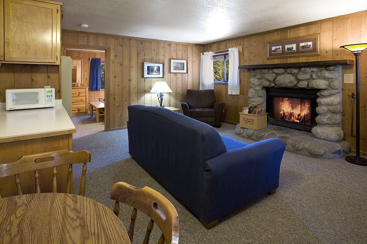 A cozy cabin interior with a stone fireplace, wooden walls, a blue sofa, a dining table, and a microwave.