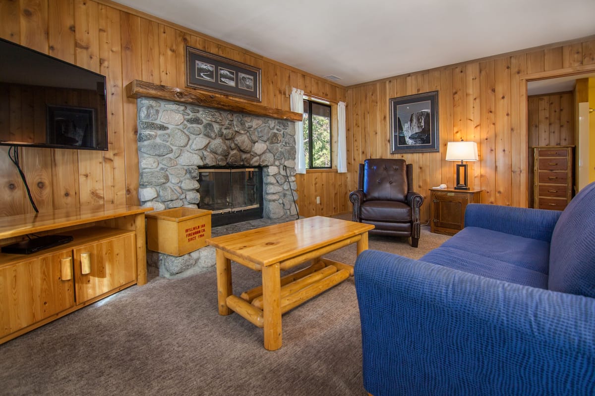 A cozy living room with wooden walls, a stone fireplace, a blue sofa, a leather armchair, and a wooden coffee table.
