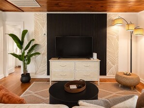 Un salon comprenant un téléviseur à écran plat, un meuble blanc avec des tiroirs, une table basse ronde, une plante en pot et un lampadaire.