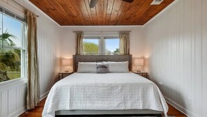 A bedroom with a wooden ceiling, a large bed with white bedding, two bedside tables, and a view of the outdoors through a window.