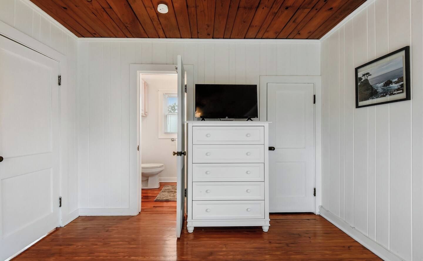 Un couloir avec une commode blanche, un téléviseur posé dessus et un tableau encadré accroché au mur.