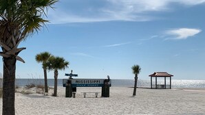 A beach with a wooden bench, a 'Welcome' sign, a gazebo, and palm trees.