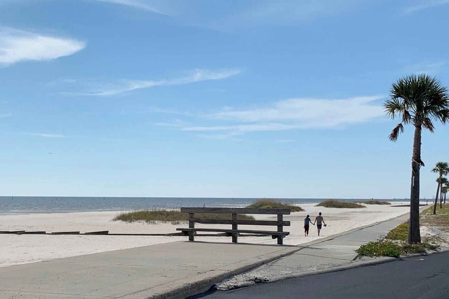 A beach with a wooden bench, a paved walkway, and palm trees.