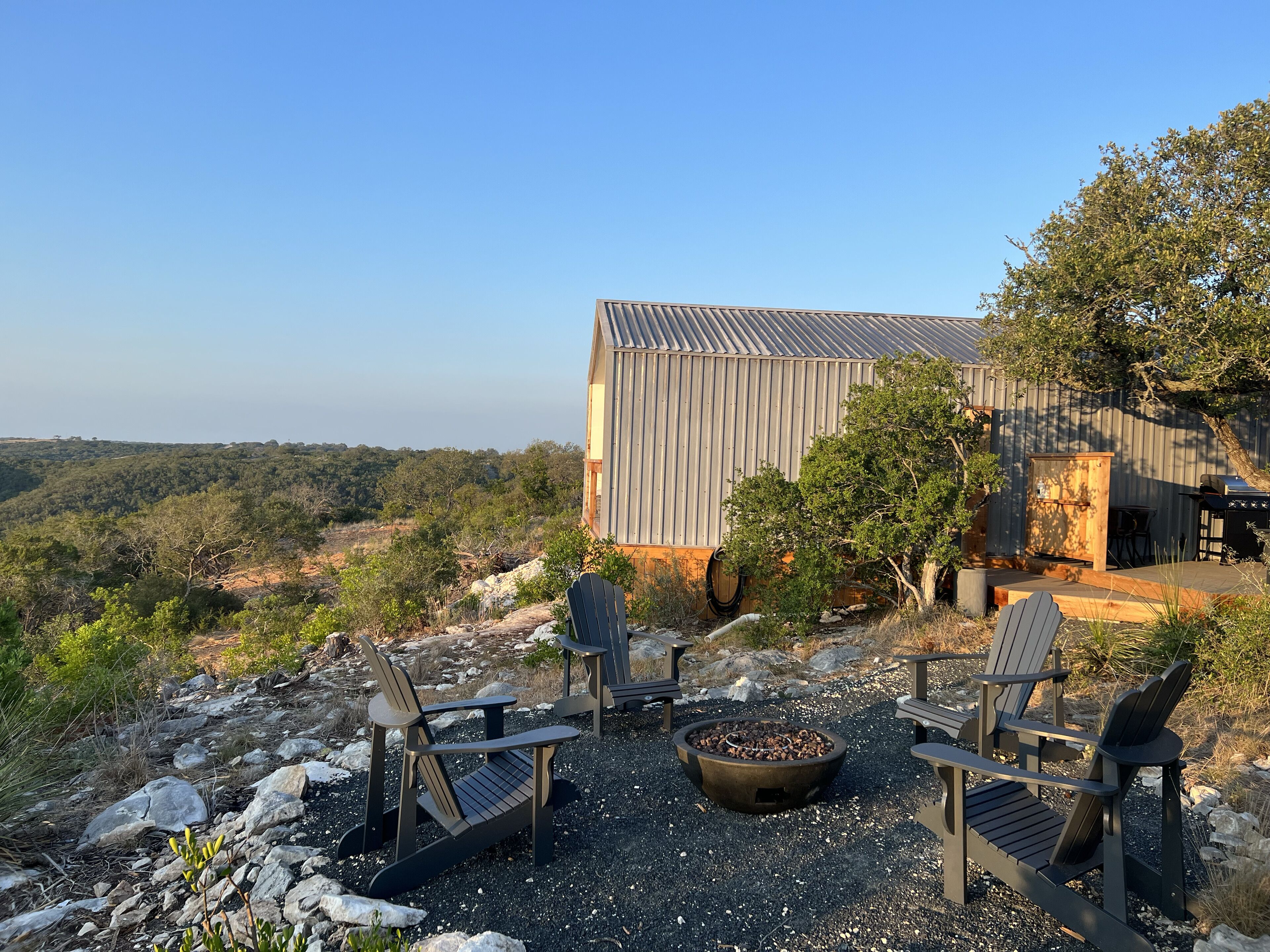 A rustic outdoor seating area with Adirondack chairs and a fire pit, overlooking a hilly landscape and a large metal barn.
