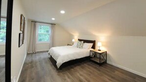 Une chambre avec un plancher en bois, un lit avec une literie blanche, deux lampes de chevet et une fenêtre avec des rideaux.