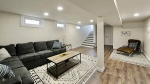 Un salon moderne avec un canapé d’angle, une table basse et un tapis à motifs. Un escalier mène à un niveau supérieur.