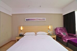 Quarto de hotel com uma cama grande, cabeceira de madeira, mesa de cabeceira com abajur, poltrona roxa e uma imagem emoldurada na parede.