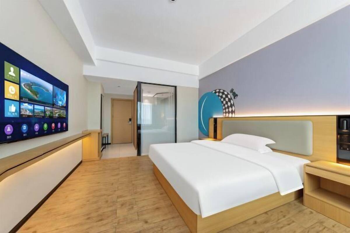 Une chambre d’hôtel moderne dotée d’une grande télévision à écran plat, d’un lit avec du linge de maison blanc, d’un plancher en bois et d’une porte en verre donnant sur la salle de bain.
