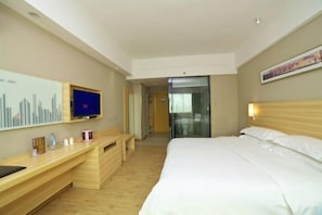 Quarto de hotel com cama, televisão sobre suporte de madeira, escrivaninha e porta para banheiro.