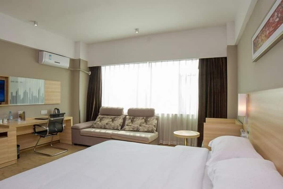 Une chambre d’hôtel avec un grand lit, un canapé avec des coussins, un bureau avec une chaise, une petite table ronde et une grande fenêtre avec des rideaux.