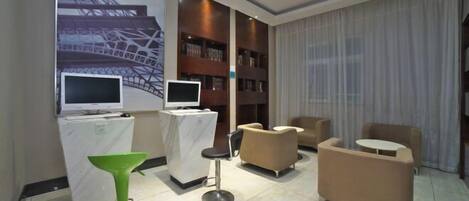 A modern hotel lobby with a large screen displaying a cityscape, a reception desk, a green bar stool, and comfortable seating areas.