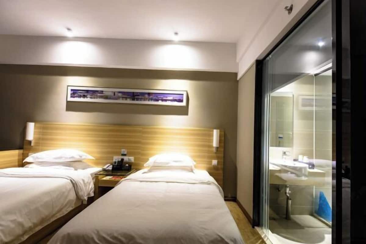 Une chambre d’hôtel avec deux lits, une table de chevet avec un téléphone, une lampe fixée au mur et une salle de bain visible à travers une porte en verre.