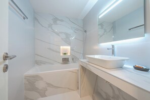A modern bathroom with a marble wall, a freestanding bathtub, a large mirror, and a wall-mounted light fixture. - KYPSELI PLATINUM (Athens)